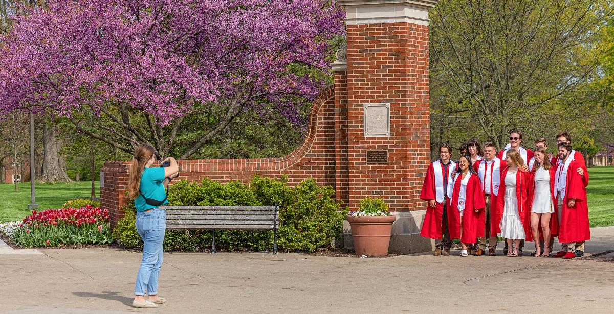 A group of Miami students posing by the Uptown gates in graduation robes for a photo