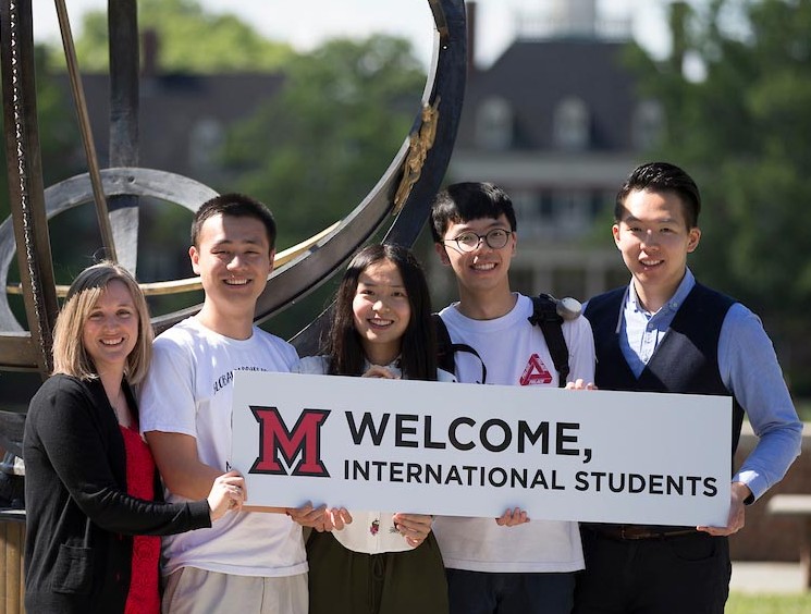 A group of international students stands in front of the Miami sundial holding a sign welcoming new international students