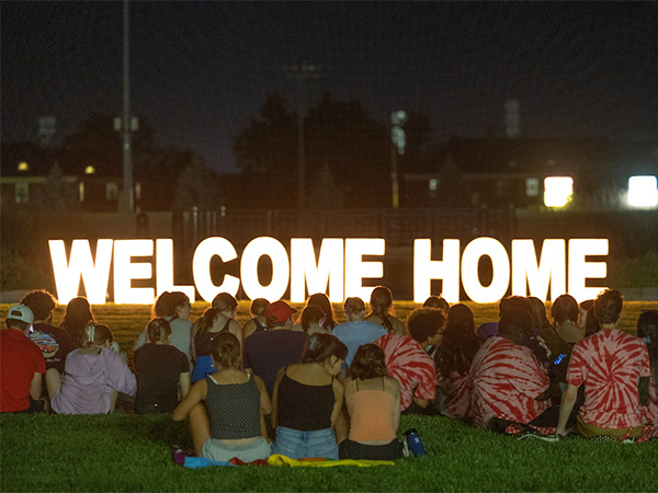 students outside at night looking at a giant light up sign that says "welcome home"